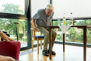 Älterer Herr im Seniorenheim setzt sich gerade hin. Der Stuhl wurde individuell für ihn nach Maß und mit erhöhten Armlehnen gefertigt.